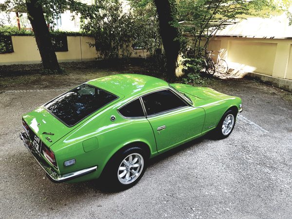 1971 Datsun 240Z in green, rear three-quarter on a gravel drive, showing chrome bumpers and eight-spoke alloy wheels