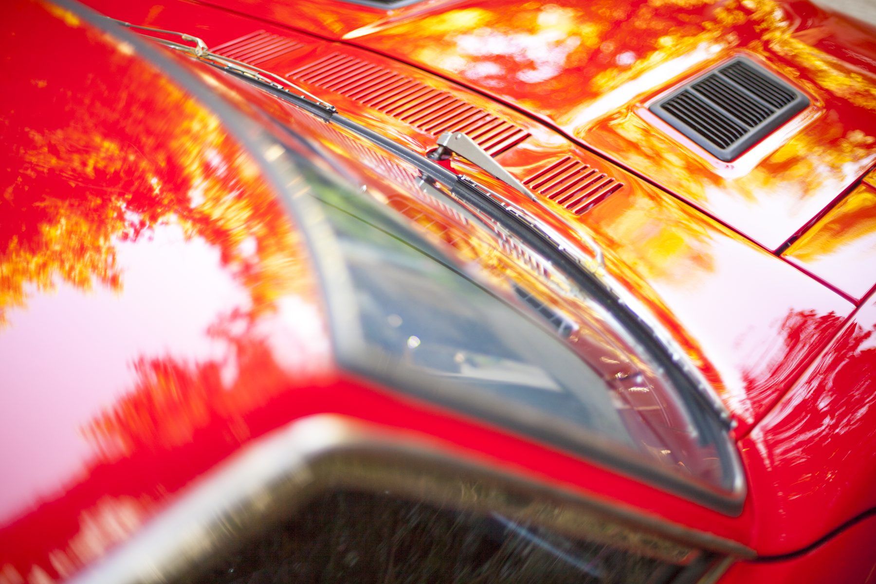 Headlight surround and bonnet of a red 1976 Datsun 280Z, foliage reflected in the paint