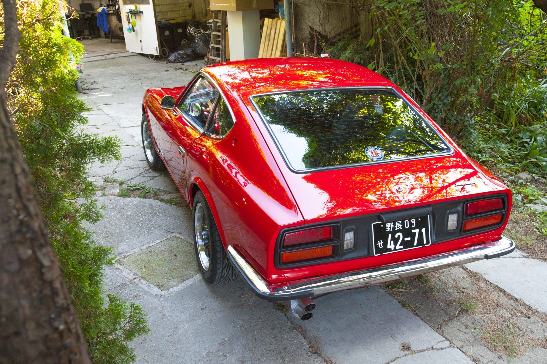 Red 1976 Datsun 280Z from the rear three-quarter outdoors, daylight showing unbroken reflections along the flank